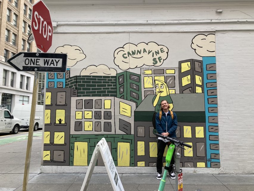 A man stands in front of a mural depicting a banana in a green shirt sitting on a rooftop smoking weed.