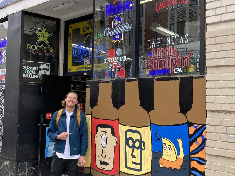A man poses next to a mural of four cartoon bottles on the outside wall of a store with prominent beer and wine signs.