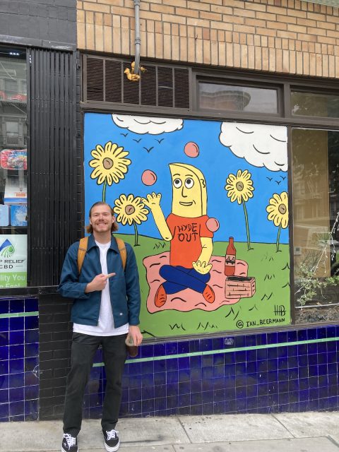 A man poses in front of a mural depicting a banana picnicking in a field of flowers and juggling.