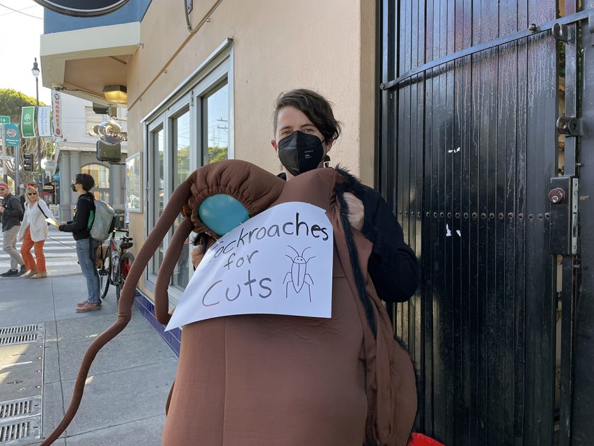 Person in a cockroach costume holding a sign that reads "Cockroaches for Cuts," standing on a city sidewalk near a building.