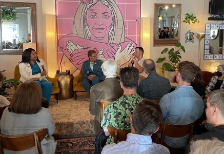 Three people engaged in a panel discussion in front of an audience. A large pink mural with a woman holding an anatomical heart is seen in the background.
