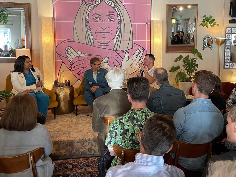 Three people engaged in a panel discussion in front of an audience. A large pink mural with a woman holding an anatomical heart is seen in the background.