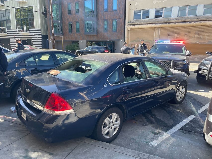 A damaged blue sedan with shattered windows is parked on the street. Police officers and a patrol car with flashing lights are present in the background, with a barricaded area surrounding the scene.