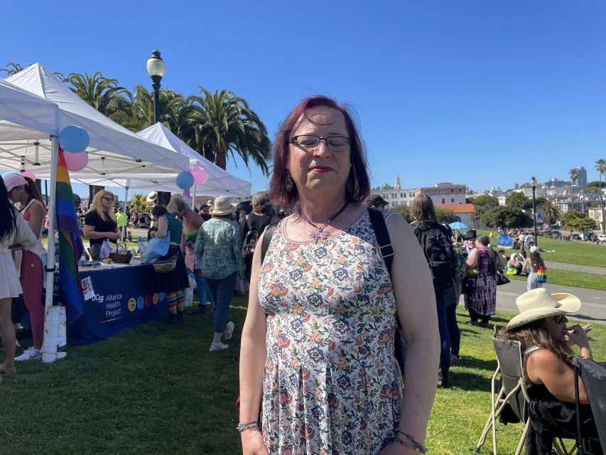 A woman poses for a photo in a park. she is wearing a dress and a dragonfly necklace