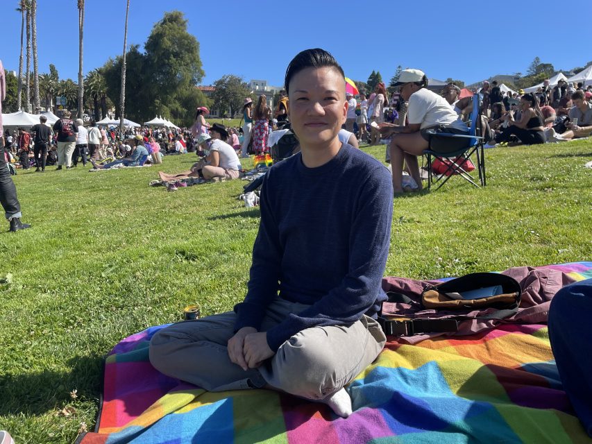 A woman sits on a rainbow picnic blanket in a park.