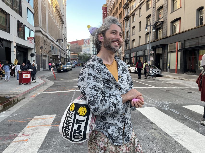 A non binary person poses in an intersection with a rucksack that says Jews for a ceasefire