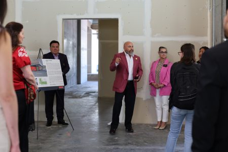 A man in a red jacket speaks to a group of people inside a construction site. A display board is near him. Other individuals, including a woman in a pink jacket, observe. The room is unfinished.