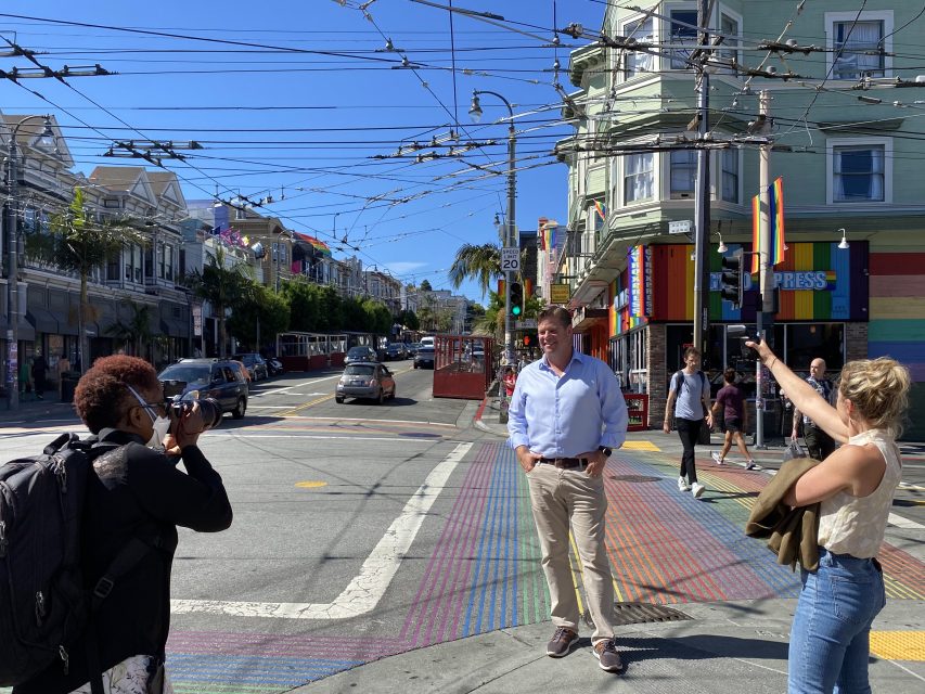 A man poses for a photo on a colorful crosswalk with two people, one taking a photo and the other pointing, at a street corner with buildings under blue sky.