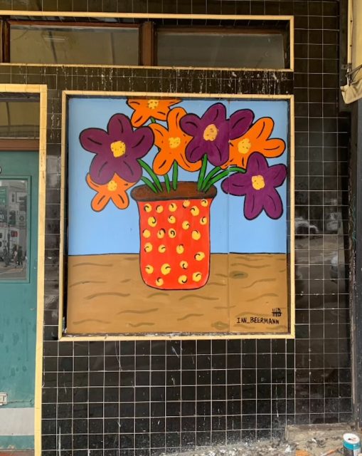 A mural depicting purple and orange flowers in a red and yellow vase on a wood table. Mural by Ian Beermann