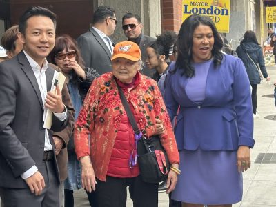 A group of people, including a woman in a purple dress and an elderly woman in an orange cap and red jacket, stand together outdoors. The background shows passersby and a "Mayor London" sign.