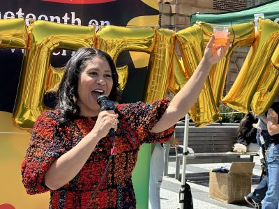 London Breed in a red and black patterned top holds a microphone and raises a drink in a celebratory gesture. Behind her are large gold balloon letters spelling "Juneteenth."