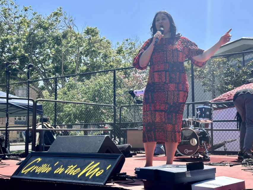 London Breed wearing a red and black patterned dress is speaking into a microphone on an outdoor stage. A sign reading "Grillin'' in the Mo'" is displayed at the front of the stage.