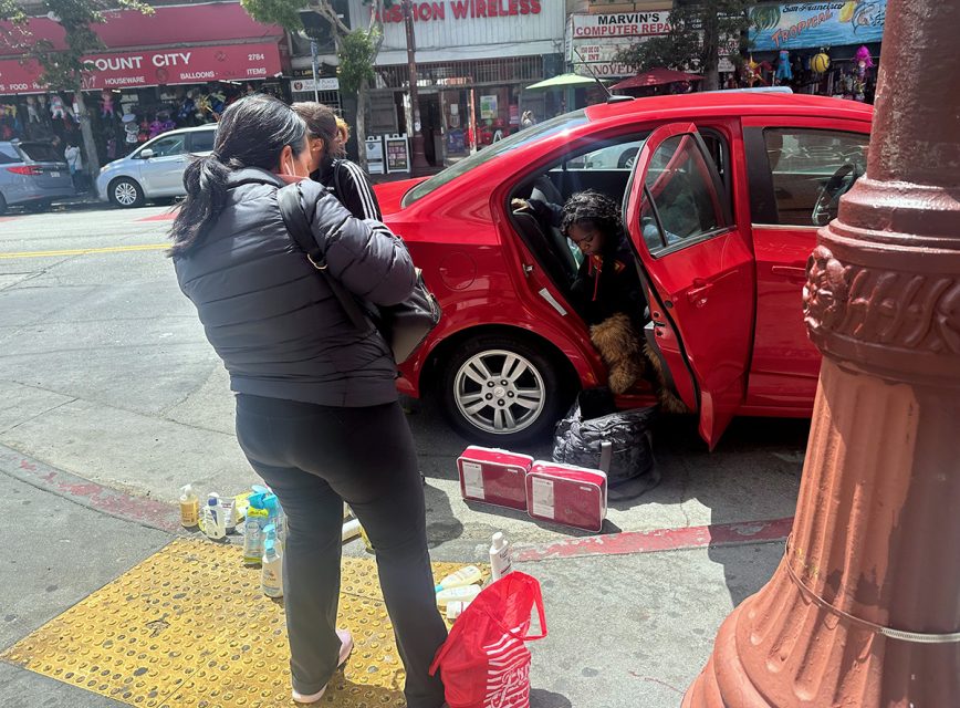Two people conversing by a red car with its doors open on a city street. One person is sitting inside the car with a dog, and the ground around them has various items placed on it.