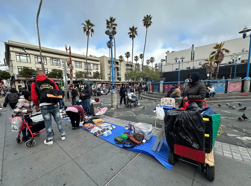 People are gathered in an outdoor market, some standing and others browsing items laid out on tarps. Palms and buildings are in the background.