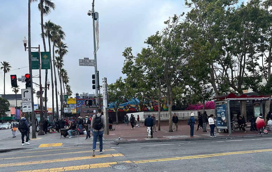 Street scene with people waiting at a bus stop and walking near a mural-covered building, surrounded by trees and palm trees. Traffic lights are visible, and the sky is overcast.