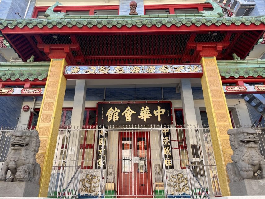 Red and green ornate entrance to a building with Chinese characters above the door, flanked by two stone lion statues and metal fencing.