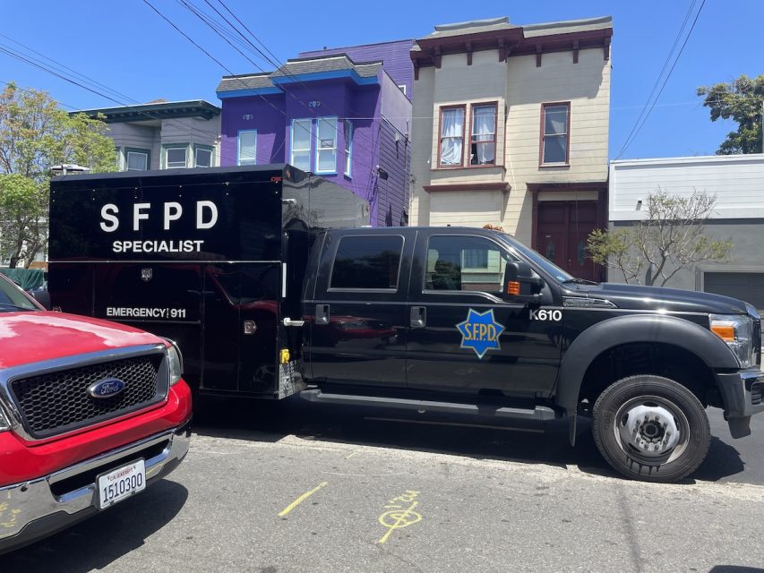 A black SFPD SWAT vehicle parked on a residential street, next to a red truck and colorful houses.