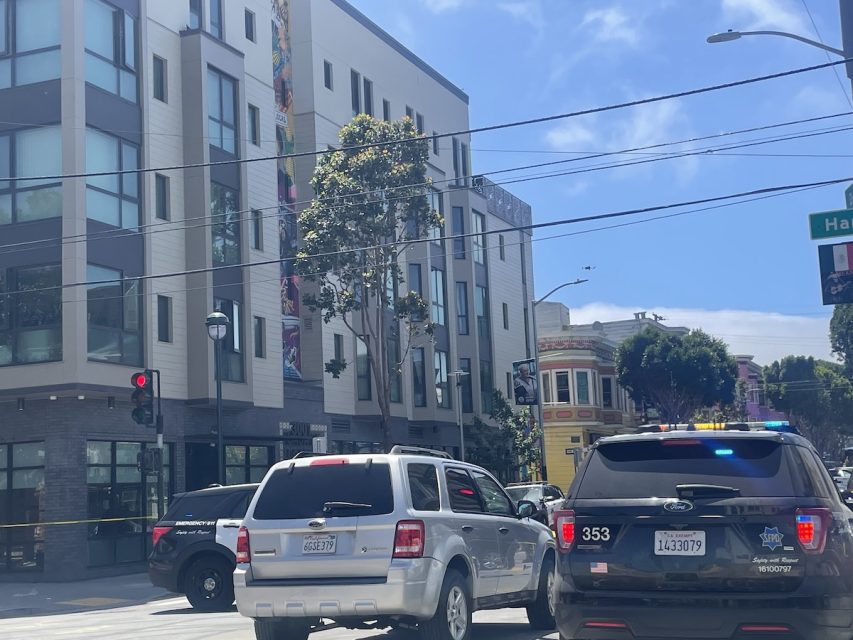 Image of a city street intersection with vehicles including police cars and an SUV. A building with large windows and a mural is in the background.