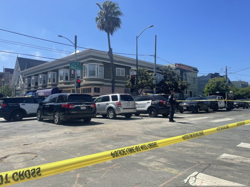 Police tape cordons off a street section near an intersection with several police vehicles and a few buildings in the background under a clear sky. A lone officer stands near the tape while a SWAT van is parked nearby, adding to the tense atmosphere.