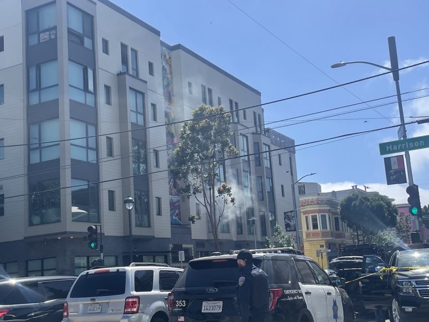 A city street scene shows a few parked SUVs, a SWAT vehicle, and a building in the background with a street sign for Harrison. Sunlight is visible with clear blue skies and minimal clouds.