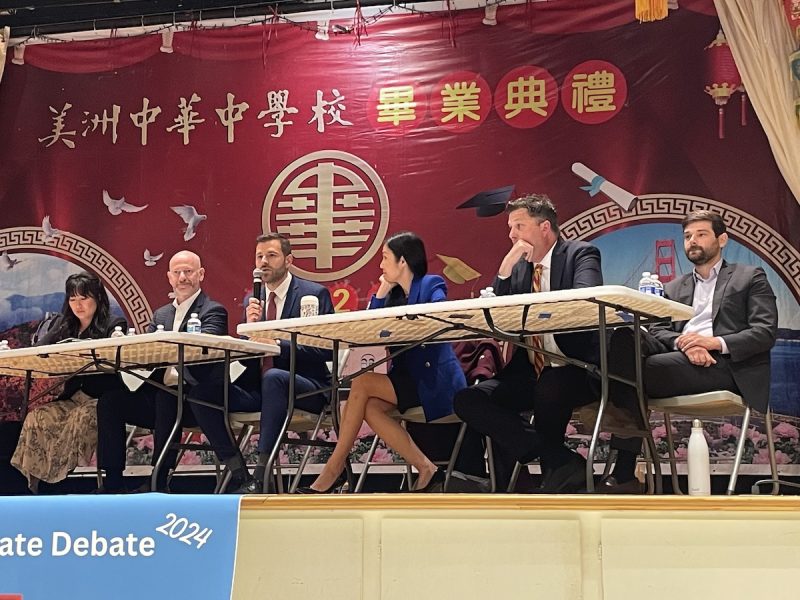 A panel of six individuals sits at a long table on a stage during a debate event at the Chinese Consolidated Benevolent Association. A red backdrop with Chinese characters and decorations is visible behind them.