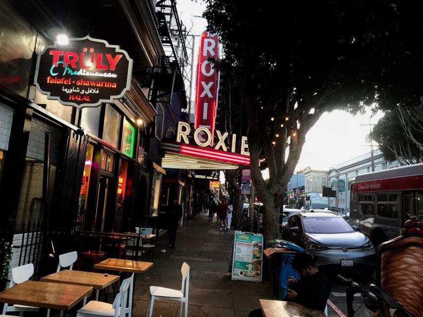 A street scene at dusk featuring the Roxie Theater with a brightly lit marquee. Outdoor seating from a nearby restaurant is visible, along with a bus and people walking on the sidewalk.