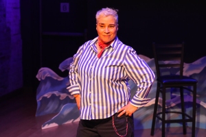 A person with short hair is standing on stage wearing a striped shirt and a red necktie. They are posed confidently with hands on hips.