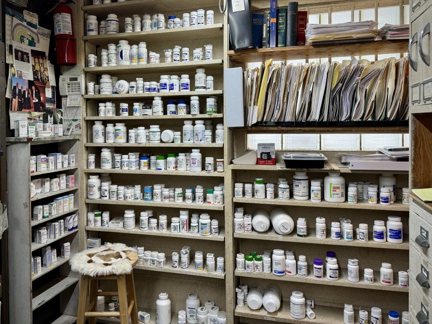 A room with shelves filled with numerous pill bottles, organized folders on top shelves, a framed photo on the wall, and a wooden stool with a fur-covered seat in front.