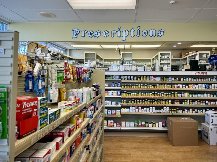 A pharmacy interior featuring shelves stocked with various over-the-counter medications and vitamins. A sign above a counter at the back reads "Prescriptions.