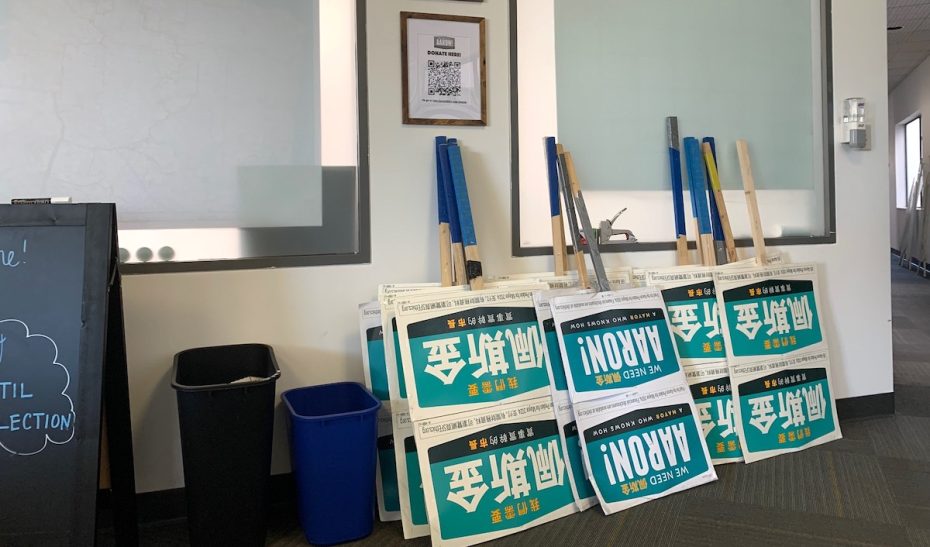 A room with campaign signs propped against the wall, a blackboard, and two recycling bins. A framed QR code is visible on the wall.