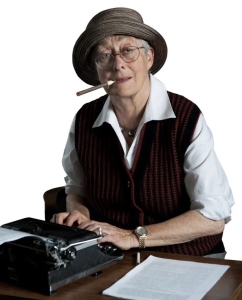 An elderly woman wearing a hat and a vest sits at a typewriter with a cigarette holder in her mouth, looking at the camera.
