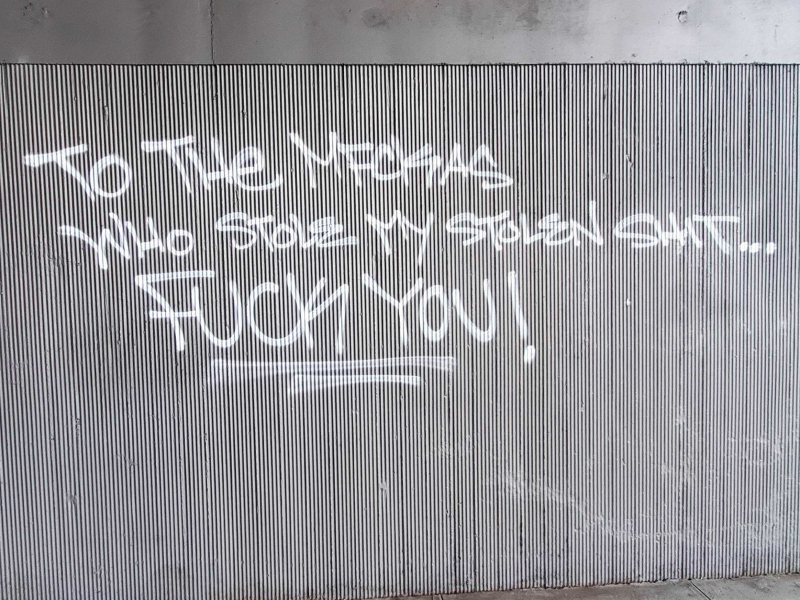 Graffiti on a ribbed metal wall reads, "To the mfckas who stole my stolen shit... FUCK YOU!