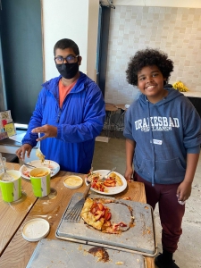 Two boys are serving and eating food at a table. One wears a blue jacket and mask, the other a gray sweatshirt. There are plates with food and empty dishes on the wooden table.