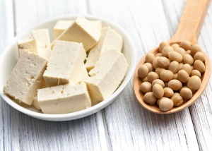 A white bowl filled with tofu cubes is placed next to a wooden spoon holding soybeans on a light wooden surface.