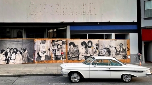 A white vintage car is parked on a street in front of a building with a large mural depicting historical black and white images of people in various scenes.