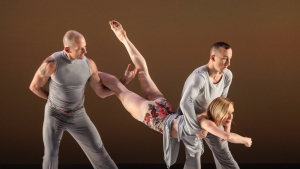Three dancers perform on stage, with two males supporting a female in a mid-air horizontal position. They wear grey outfits, and the background is a warm gradient.