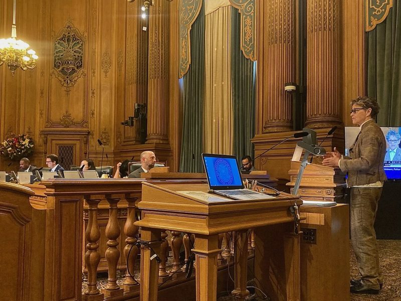 A person is speaking at a podium in a formal meeting room with ornate wood paneling. Several people are seated at desks with microphones, looking towards the speaker. A laptop and display are visible.
