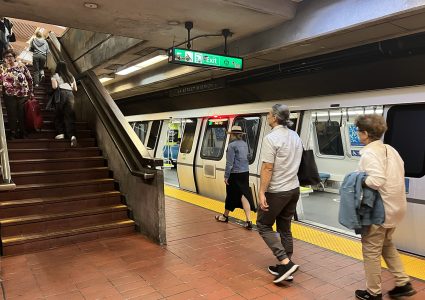 Commuters are seen boarding and disembarking a subway train, while others ascend a flight of stairs within the station. An "Exit" sign hangs overhead.