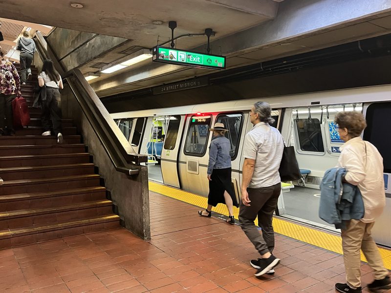 Commuters are seen boarding and disembarking a subway train, while others ascend a flight of stairs within the station. An "Exit" sign hangs overhead.