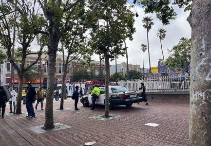 A police car is parked on a brick pathway near some trees. People are walking around, and two officers appear to be interacting with pedestrians. Buildings and a mural are visible in the background.