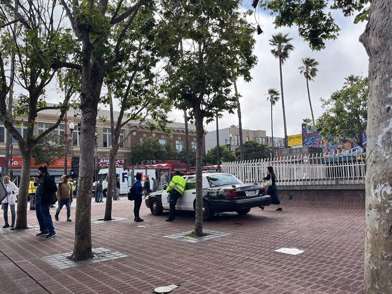 A police car is parked on a brick pathway near some trees. People are walking around, and two officers appear to be interacting with pedestrians. Buildings and a mural are visible in the background.