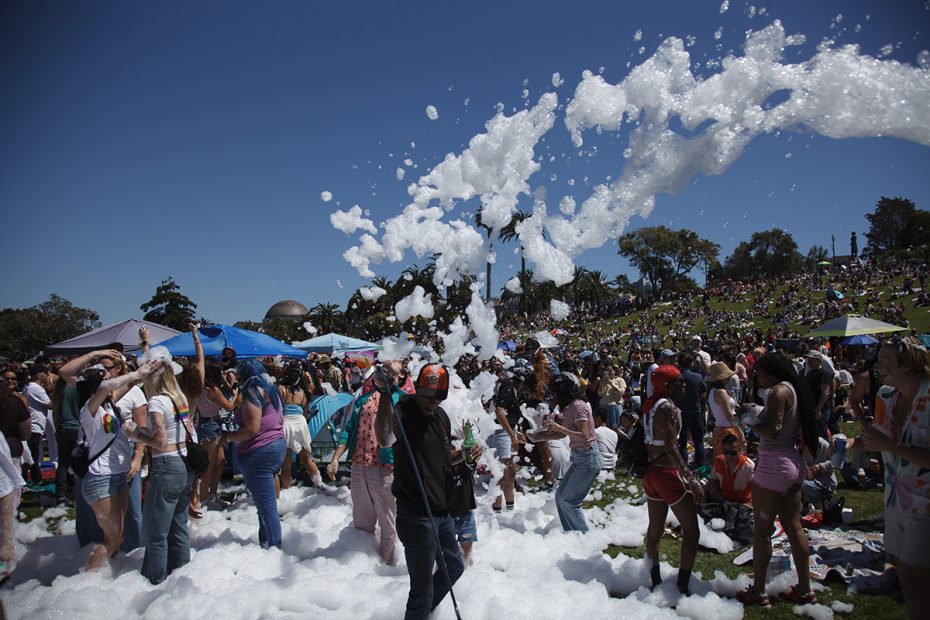 A large crowd of people at an outdoor event play in foam spray under a clear blue sky, with many others gathered on a grassy hill in the background.