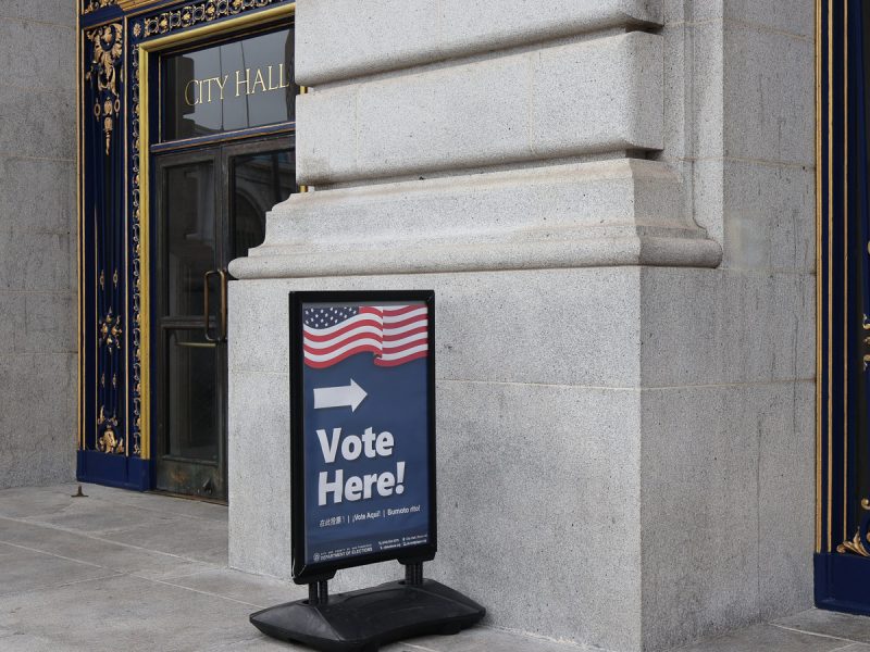 A sign with the American flag and "Vote Here!" points toward the entrance of a city hall building.