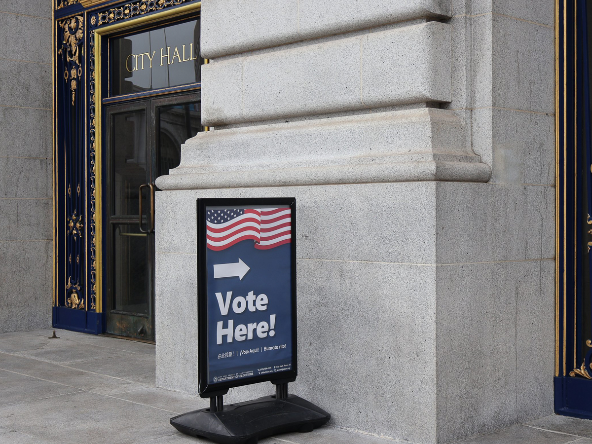 A sign with the American flag and "Vote Here!" points toward the entrance of a city hall building.