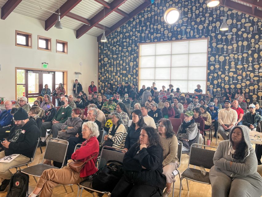 在一间窗户很高，墙上挂满了各种桨的房间里，一大群人正坐着聚精会神地听着会议。