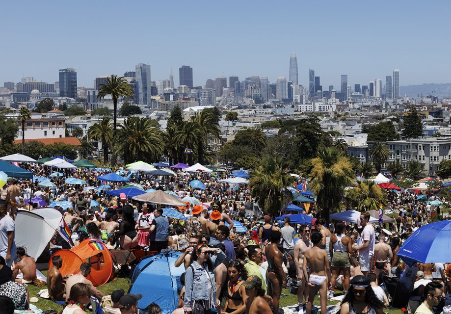 A large crowd gathers in a park on a sunny day, with numerous umbrellas and tents. The city skyline is visible in the background.