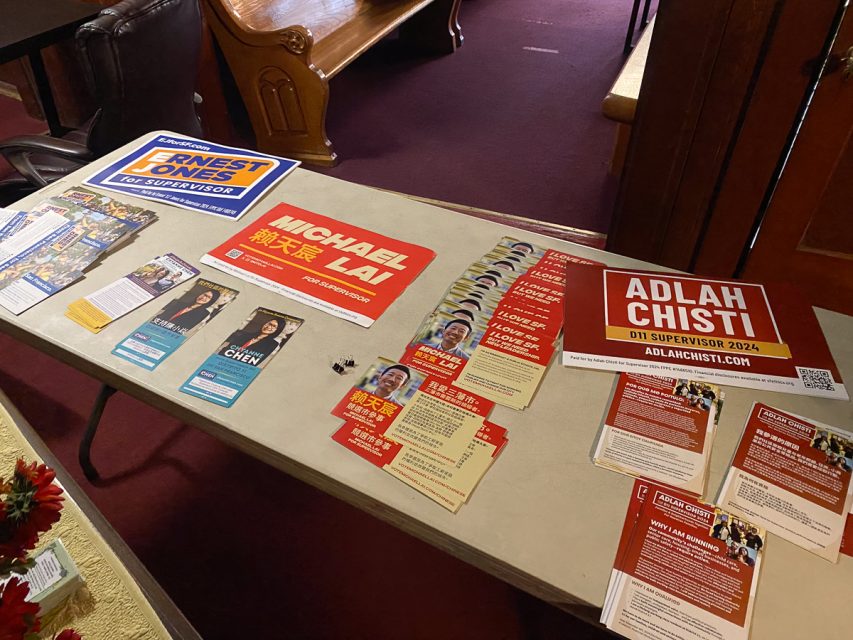 A table with campaign materials for several candidates, including flyers, brochures, and posters for District 11 candidates Ernest Jones, Michael Lai, Chyanne Chen, and Adlah Chisti, displayed on a beige tablecloth.