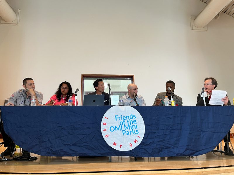 A group of six individuals sits behind a table with a blue cloth that reads "Friends of the Ohlone Mini Parks." They appear to be speaking or preparing to speak in a panel discussion setting.