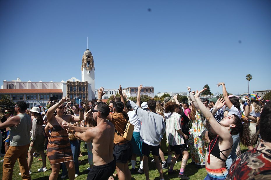 A large group of people gather outdoors in front of a historic building under a clear sky, many with arms raised or dancing, indicating a festive or celebratory event.