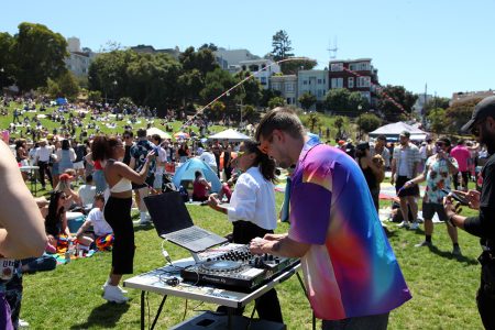 A DJ wearing a colorful shirt plays music from a laptop and mixer in a park filled with people enjoying various activities on a sunny day.